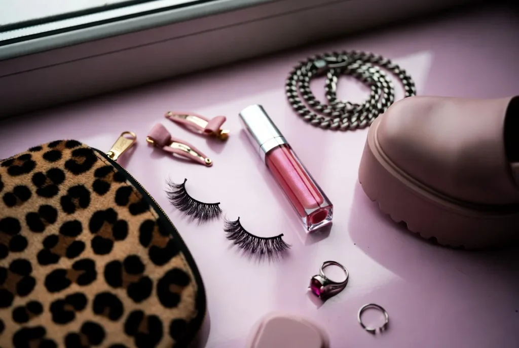 Configuração plana de itens essenciais de gyaru para iniciantes, incluindo cílios postiços, lábio brilhante, laços de cabelo, bolsa de leopardo, cinto de corrente e sapato de plataforma em superfície rosa