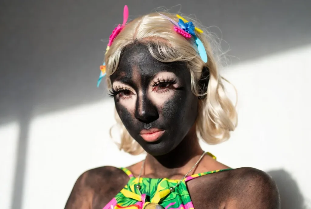 Young woman in modern yamanba gyaru recreation with extreme dark tan, platinum blonde hair, large white face accents and tropical outfit, Egg magazine inspired studio portrait